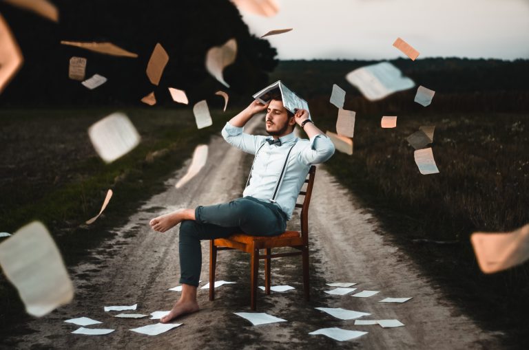 Hombre sentado en una silla de madera sosteniendo un libro abierto sobre su cabeza y cayendo hojas de papel