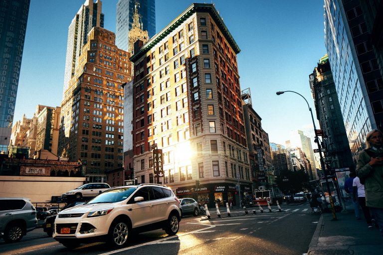 Centro urbano con edificios grandes, pegando el sol a un costado de ellos, avenida con carros circulando.